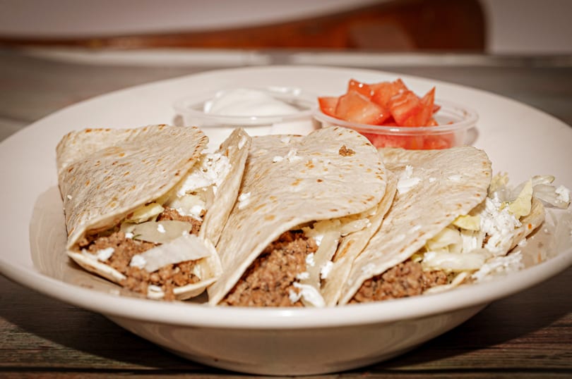 Three soft tacos filled with ground meat, cheese, and lettuce, served with sour cream and diced tomatoes.