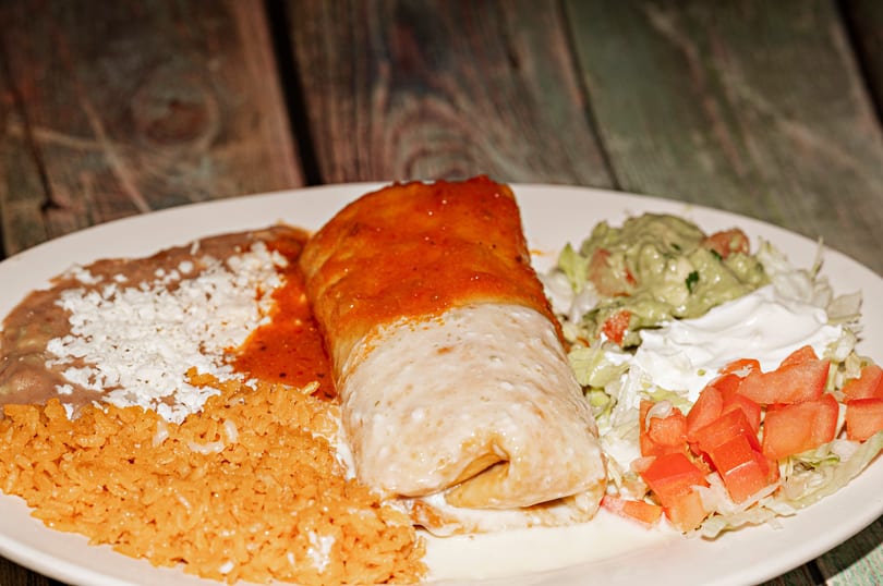 A burrito covered in red and white sauce, with rice, beans, guacamole, and salad on a white plate.