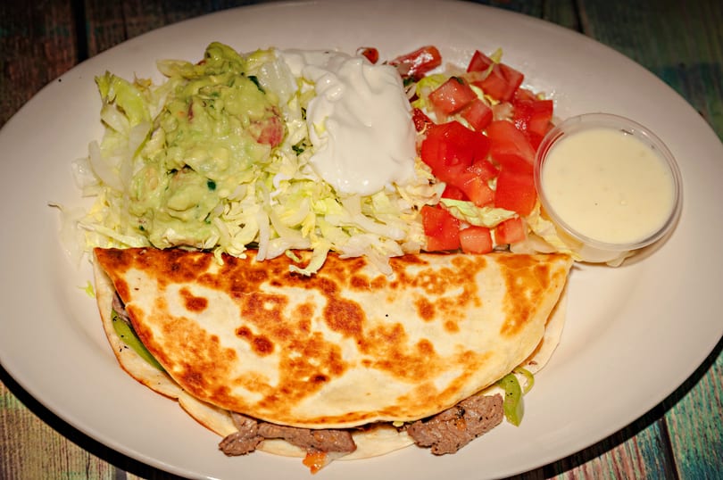A beef quesadilla with guacamole, sour cream, diced tomatoes, shredded lettuce, and a side of sauce.